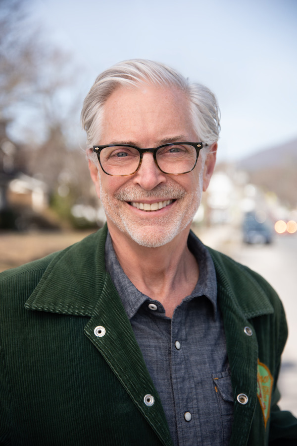 Bennet Ratcliff is a white man with gray hair wearing glasses with close shaved facial hair wearing a green jacket with lapels and a gray shirt underneath. He is smiling
